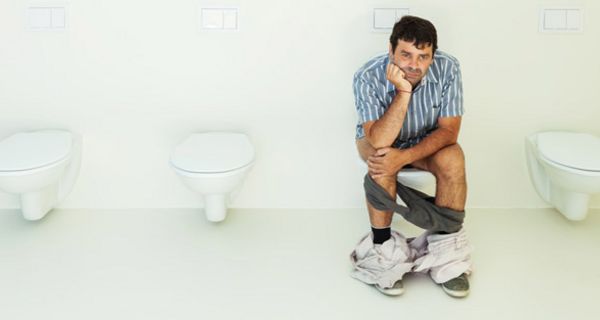 Mann, ca. Anfang/Mitte 40, sitzt mit miesepetrigem Gesicht auf einer Toilette, heruntergelassene grauer Slip, helle Hose um die Fußknöchel, eine Hand aufgestützt, die andere über den Knien