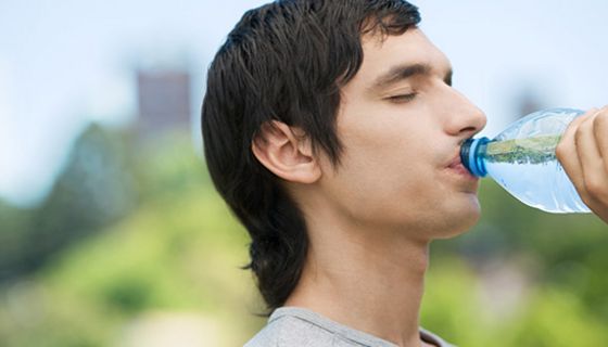 Junger Mann mit dunklen Haaren (Profilfoto vor unscharfem Naturhintergrund) trinkt aus einer Wasserflasche