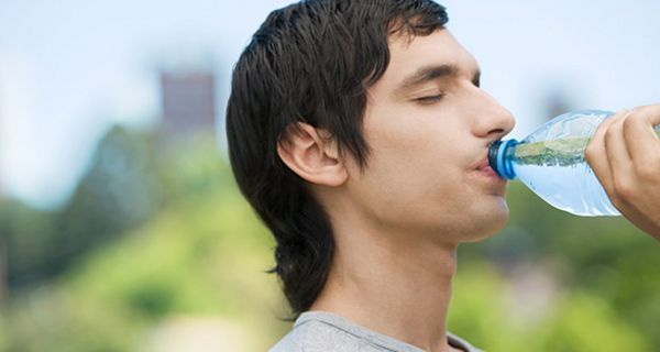Junger Mann mit dunklen Haaren (Profilfoto vor unscharfem Naturhintergrund) trinkt aus einer Wasserflasche