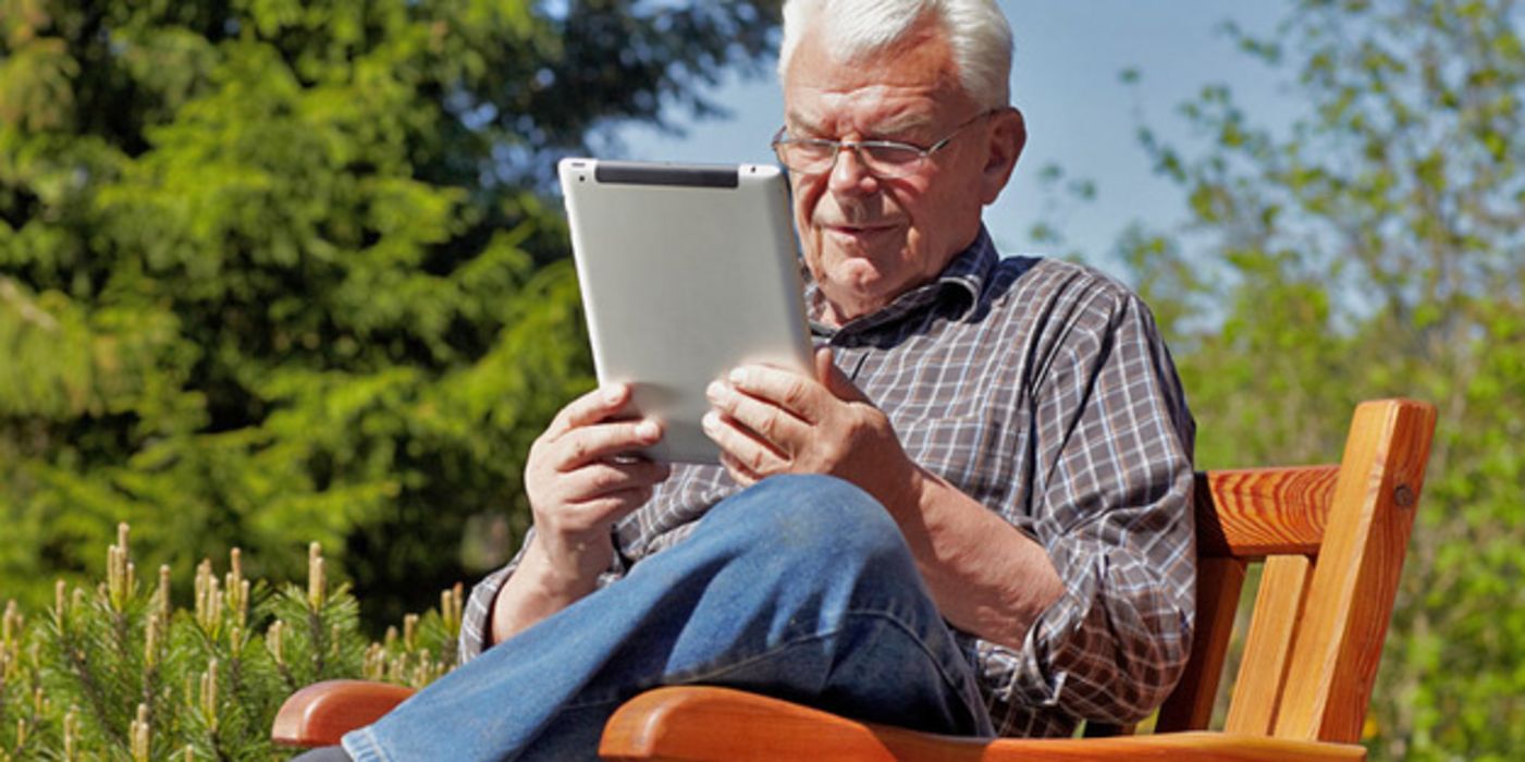 Senior sitzt im Garten und liest in einem Digitalbuch