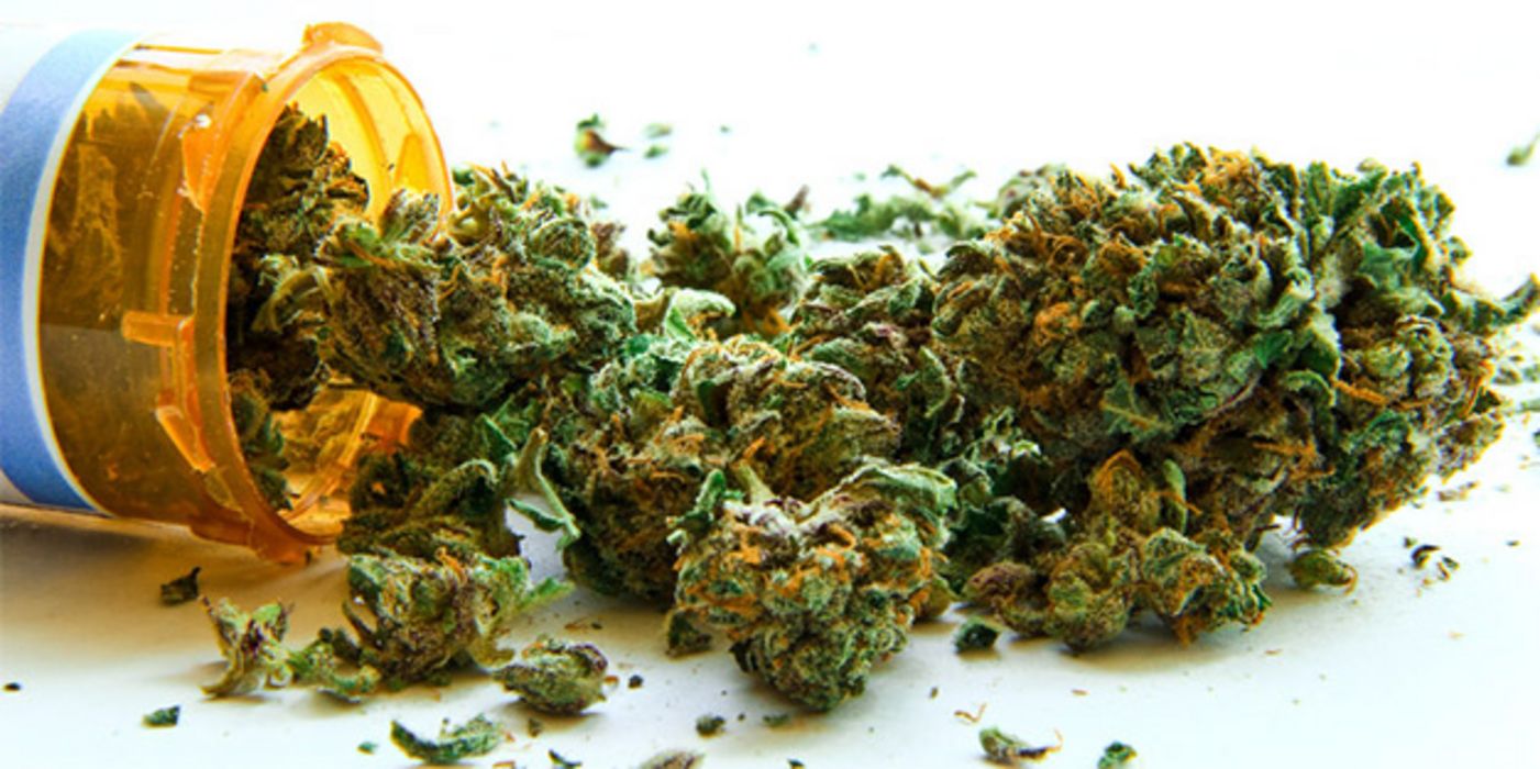 Getrocknete Cannabisblätter auf weißem Untergrund, am linken Bildrand liegendes Arzneidöschen (braun, Plastik), aus dem sie geschüttet worden sind