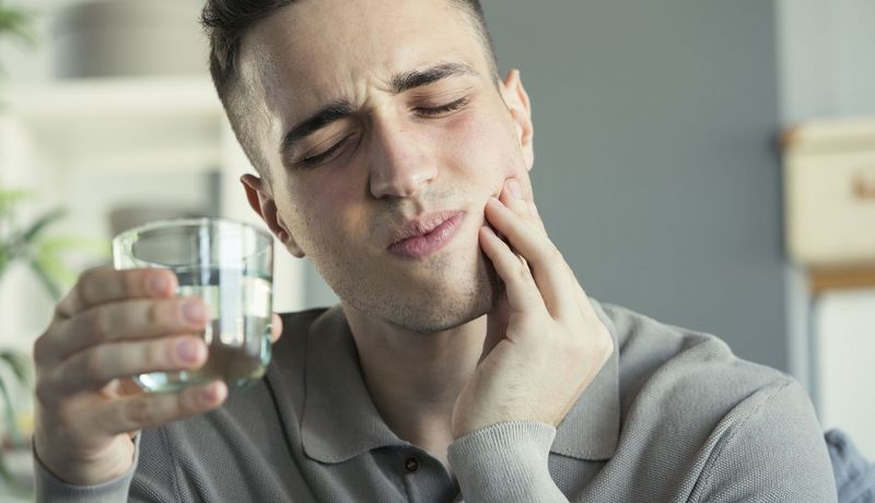 Ein junger Mann hält sich mit schmerzverzerrtem Gesicht die Wange, in der anderen Hand ein Wasserglas.