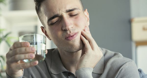 Ein junger Mann hält sich mit schmerzverzerrtem Gesicht die Wange, in der anderen Hand ein Wasserglas.