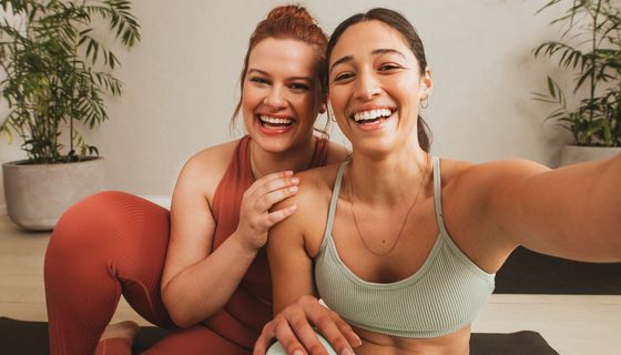 Zwei Freundinnen in Yoga-Outfit, schauen in die Kamera und lachen.