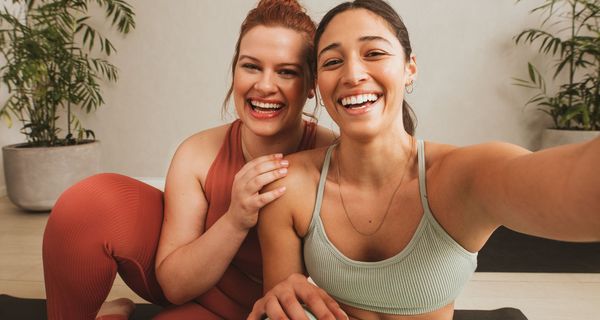 Zwei Freundinnen in Yoga-Outfit, schauen in die Kamera und lachen.