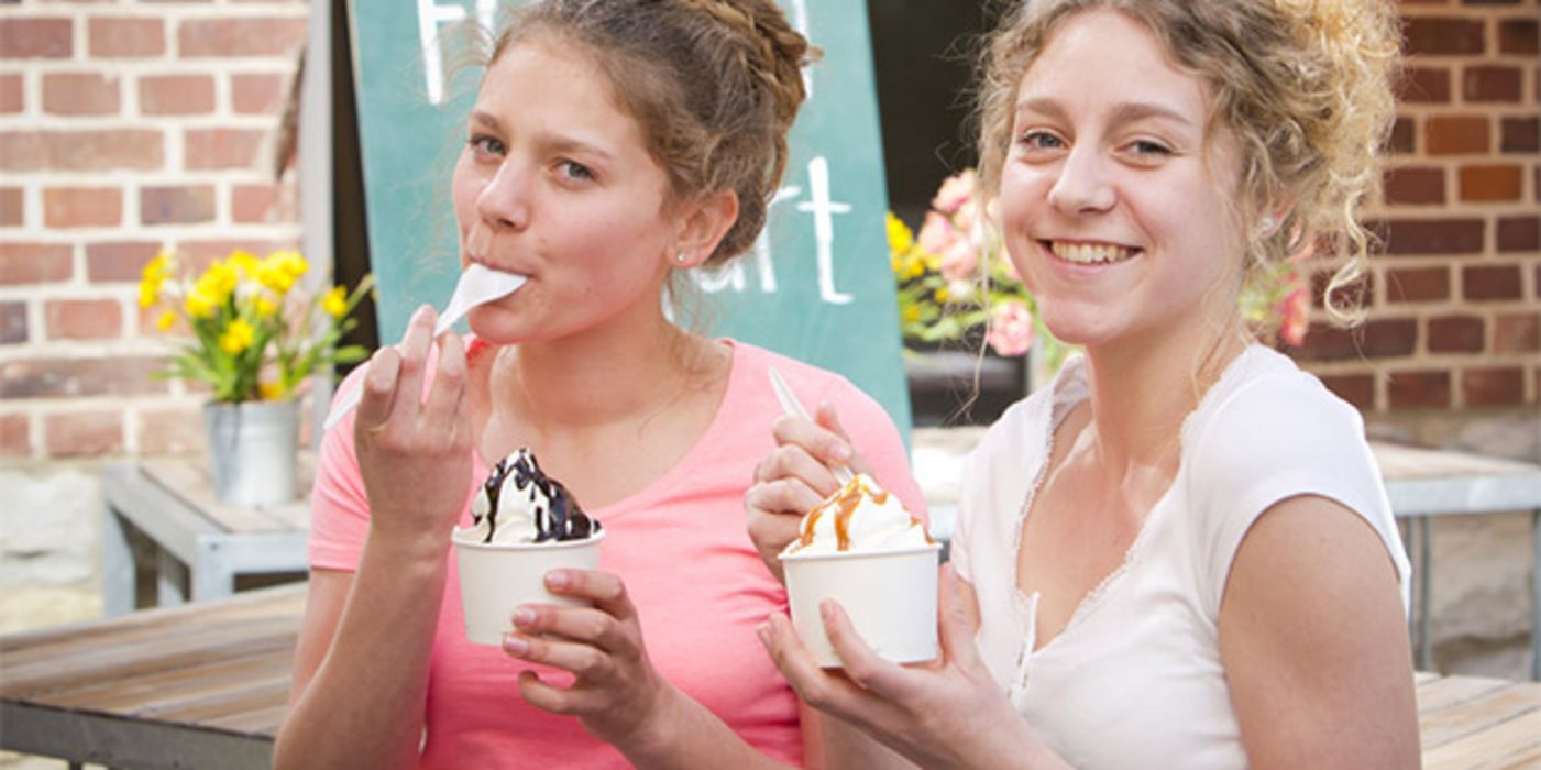 Zwei sehr junge, natürliche Frauen draußen beim Essen von Frozen-Yoghurt-Eis im Pappbecher