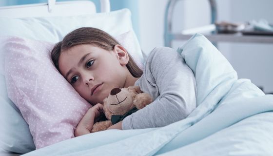 Ein trauriges Mädchen liegt im Krankenhaus, einen Teddybär im Arm.