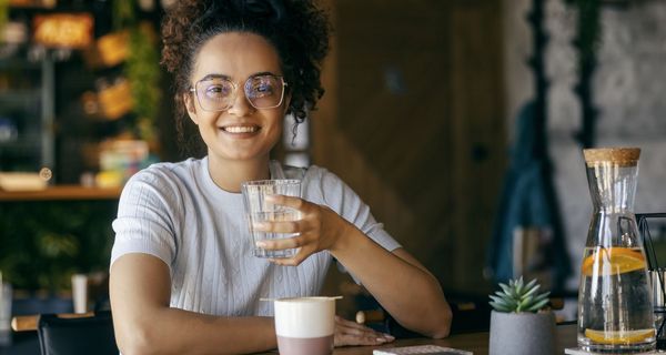 Junge Frau trinkt ein Glas Wasser, vor ihr steht eine Tasse mit einem Heißgetränk.