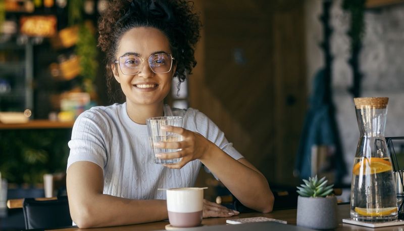 Junge Frau trinkt ein Glas Wasser, vor ihr steht eine Tasse mit einem Heißgetränk.