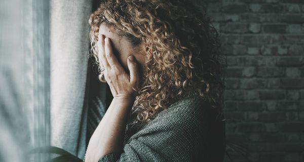 Frau steht vor dem Fenster und bedeckt ihr Gesicht mit ihren Händen.