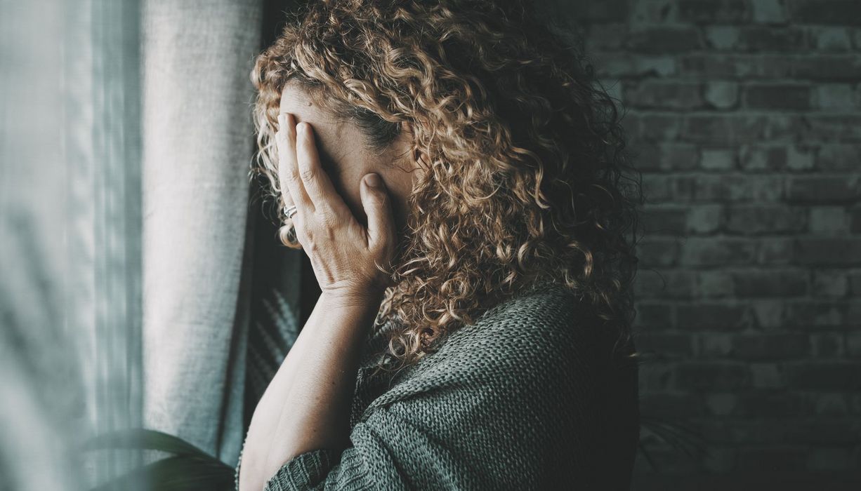 Frau steht vor dem Fenster und bedeckt ihr Gesicht mit ihren Händen.