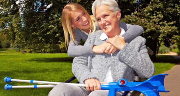 Parkszene: Attraktive, grauhaarige Frau im Rollstuhl mit 2 Krücken über die Knie gelegt, wird von hinten von blonder Tochter umarmt. 