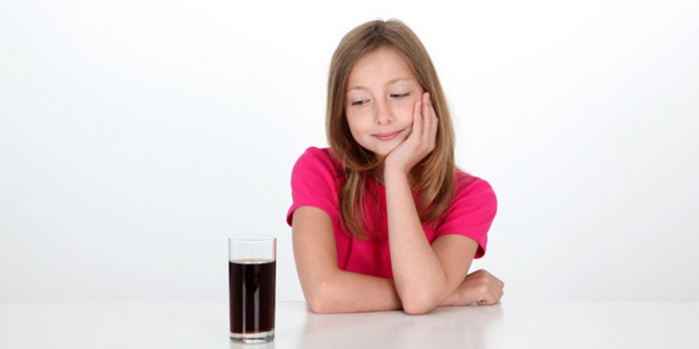 Mädchen mit langen, braunen Haaren, ca. 10/11 Jahre alt, in Kurzarmpoloshirt, Kopf in linke Hand gestützt, schaut sehnsüchtig auf ein Glas Cola