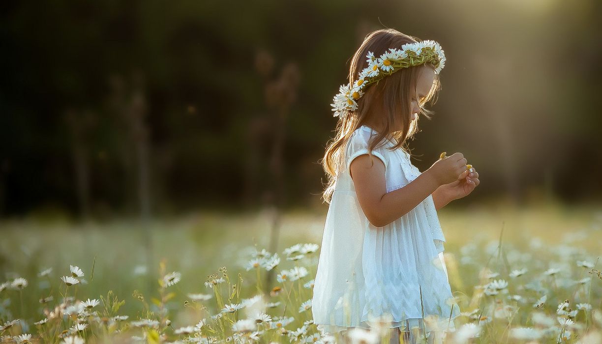 Junges Mädchen steht in einer Blumenwiese und betrachtet eine Blume.