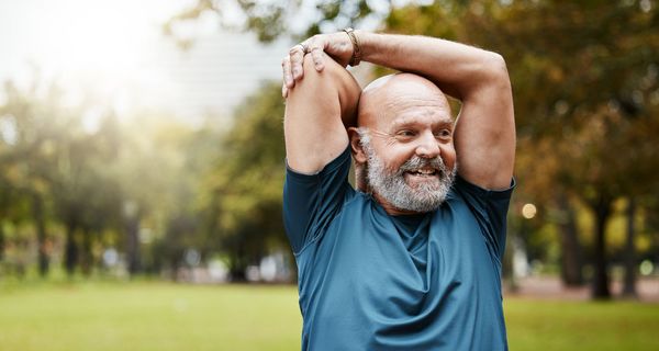 Älterer Herr mit grauem Bart und Sportshirt dehnt sich im Park.