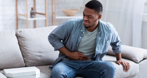 Ein junger Mann sitzt auf dem Sofa und hält sich den Bauch. Neben ihm liegt eine Pizza, die er leicht angeekelt betrachtet.