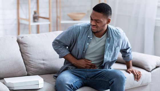 Ein junger Mann sitzt auf dem Sofa und hält sich den Bauch. Neben ihm liegt eine Pizza, die er leicht angeekelt betrachtet.