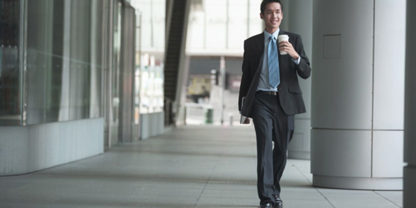 Jüngerer Asiate im Business-Anzug, blauer Schlips, lächelnd, Coffee to go in der Hand, läuft an einem Bürogebäude vorbei