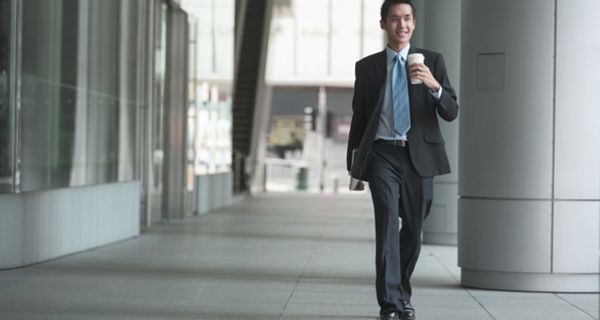 Jüngerer Asiate im Business-Anzug, blauer Schlips, lächelnd, Coffee to go in der Hand, läuft an einem Bürogebäude vorbei