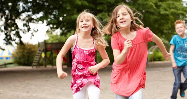 Schulhof: 2 Mädchen, Sommertops (rot, rosa), weiße Jeans, ca. 10 Jahre alt, rennen mit fliegenden Haaren Richtung Kamera; im Hintergrund ein Junge in Jeans und hellblauem Shirt