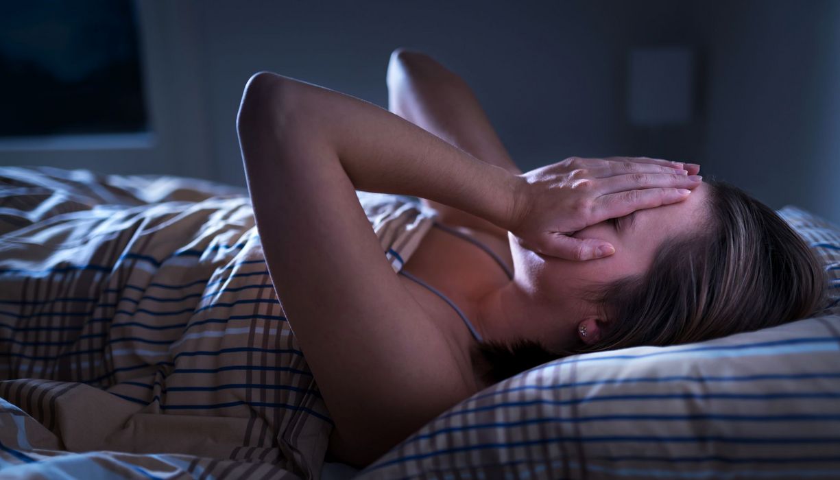 Frau, liegt im Bett und schlägt ihre Hände auf das Gesicht.