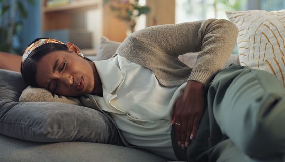 Eine junge Frau krümmt sich mit schmerzverzerrtem Gesicht  auf dem Sofa. Ihr Arm liegt auf ihrem Bauch, knapp über den Hüften. 