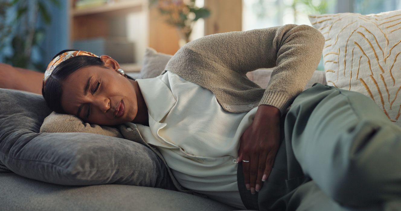 Eine junge Frau krümmt sich mit schmerzverzerrtem Gesicht  auf dem Sofa. Ihr Arm liegt auf ihrem Bauch, knapp über den Hüften. 