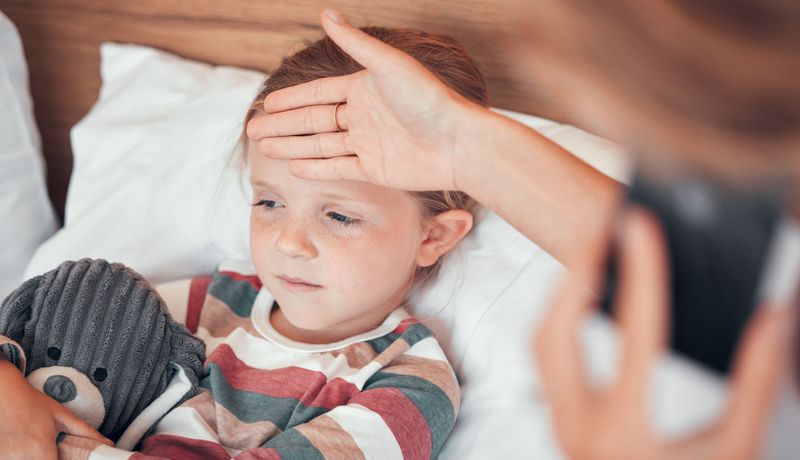 Ein kleines Mädchen liegt im Bett mit einem Kuscheltier in der Hand. Eine Person, die nicht erkennbar ist hält ihre Hand an die Stirn.