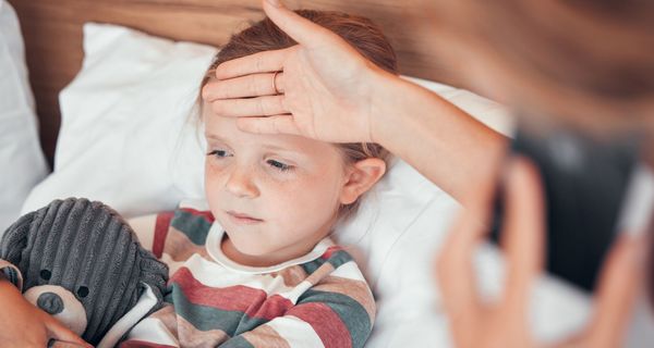 Ein kleines Mädchen liegt im Bett mit einem Kuscheltier in der Hand. Eine Person, die nicht erkennbar ist hält ihre Hand an die Stirn.
