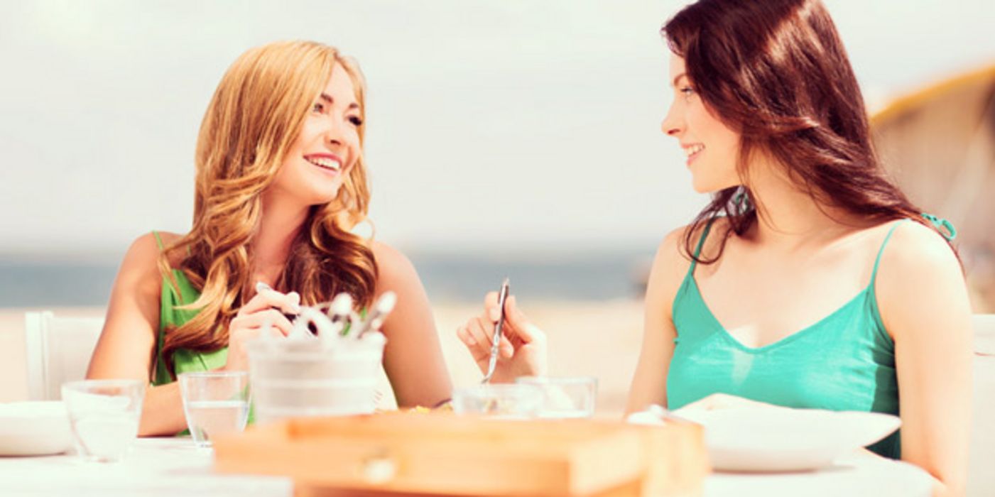 Sommerstimmung: Zwei junge Frauen sitzen im Freien im Café, essen etwas und unterhalten sich lachend dabei