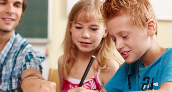 Ein Junge und ein Mädchen sitzen im Klassenraum und bekommen von einem jungen Lehrer etwas erklärt