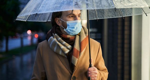 Mann mit Mund-Nasen-Schutz hält Regenschirm.