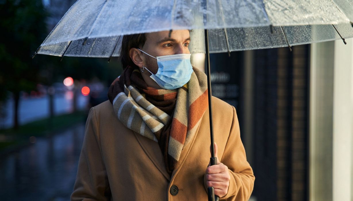 Mann mit Mund-Nasen-Schutz hält Regenschirm.
