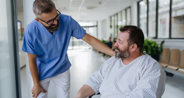 Übergewichtiger Mann im Rollstuhl, daneben steht ein Arzt und legt ihm eine Hand auf die Schulter.