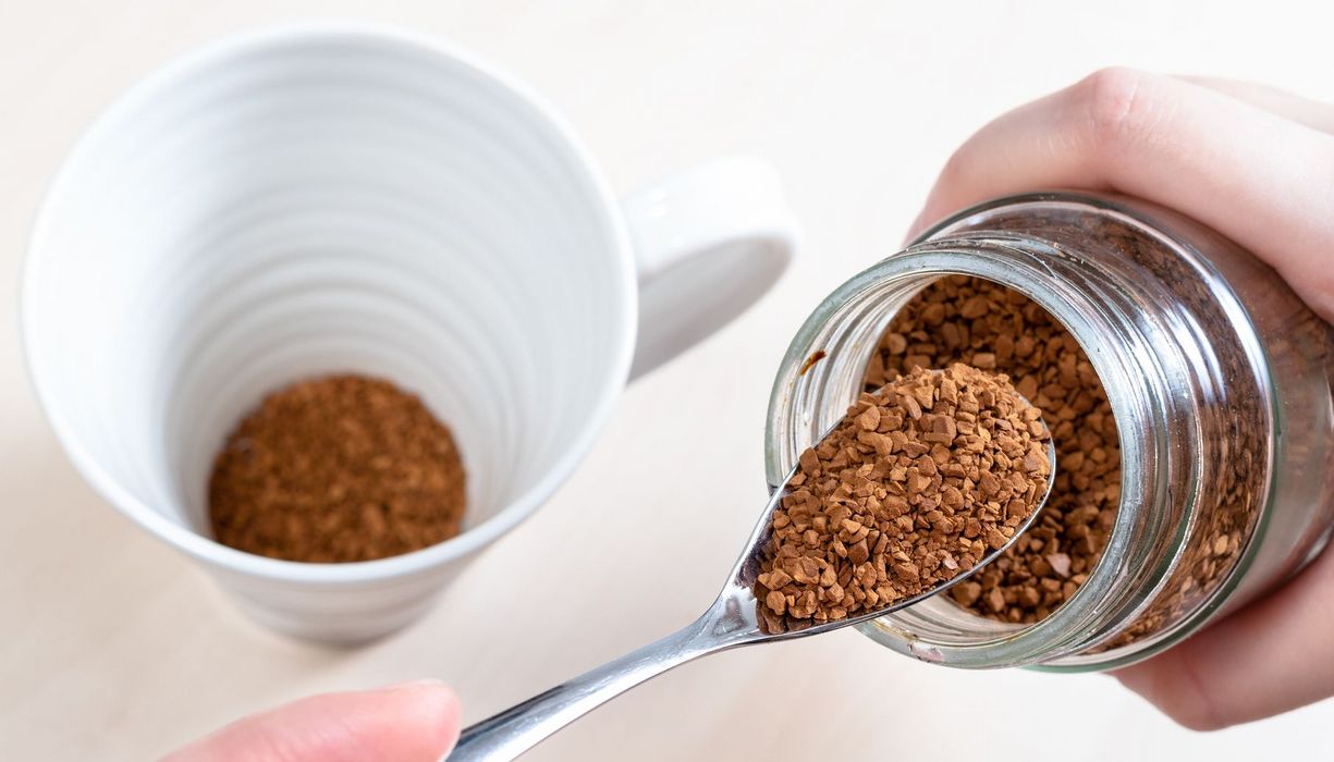 Jemand löffelt Instant-Kaffee-Pulver in eine Tasse. Nur die Hände sind zu sehen.