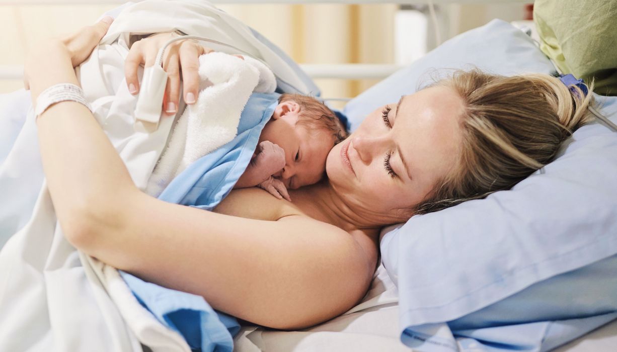 Baby liegt im Krankenhaus auf der Brust der Mutter, beide nackt und zugedeckt.