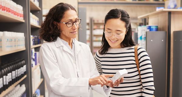 Eine Apothekerin hält ein Arzneimittel in der Hand und spricht mit einer zweiten Frau, die ebenfalls das Arzneimittel bertrachtet.