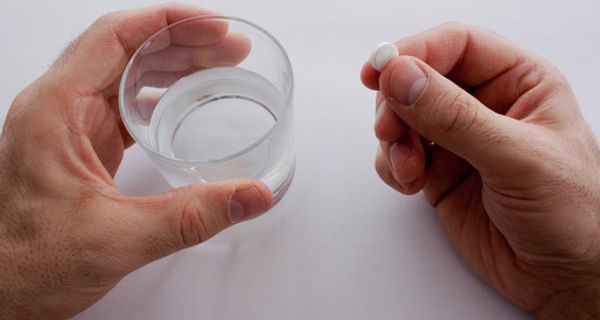 Zwei Hände auf weißerm Untergrund, von oben fotografiert: linke Hand hält Wasserglas, rechte eine längliche Tablette mit einer Einkerbung in der Mitte