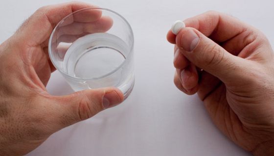 Zwei Hände auf weißerm Untergrund, von oben fotografiert: linke Hand hält Wasserglas, rechte eine längliche Tablette mit einer Einkerbung in der Mitte