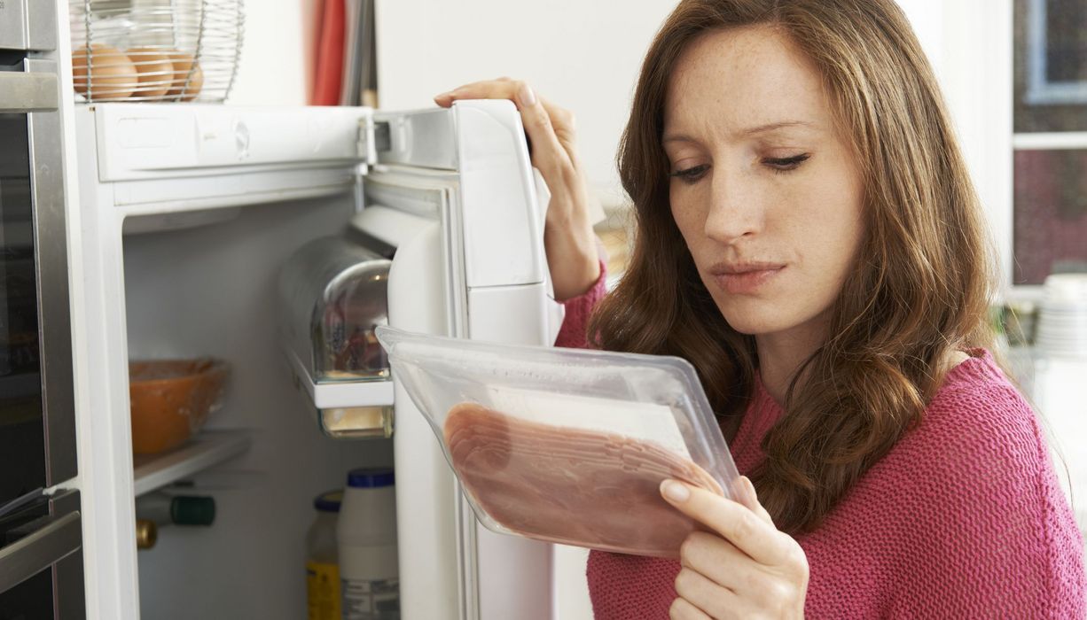 Frau betrachtet eine Packung Schinken, steht vor dem Kühlschrank.