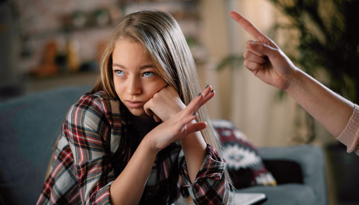 Junges Mädchen schaut genervt zur Seite, während ihre Mutter sie mit dem erhobenen Finger streng zurecht weist. 
