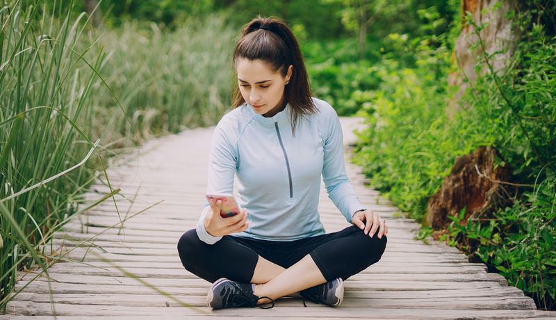 Junge Frau sitzt mit einem unzufriedenen Gesichtsausdruck in Sportkleidung auf einem Holzweg und blickt auf ihr Smartphone.