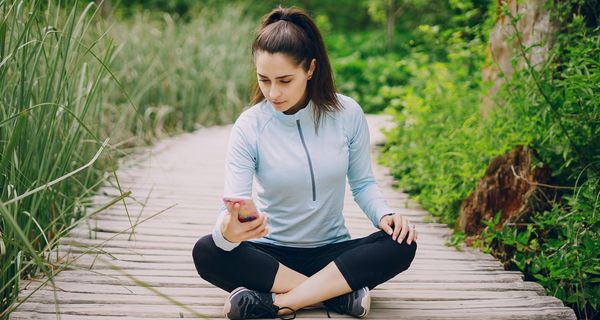 Junge Frau sitzt mit einem unzufriedenen Gesichtsausdruck in Sportkleidung auf einem Holzweg und blickt auf ihr Smartphone.