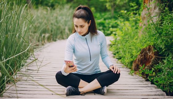 Junge Frau sitzt mit einem unzufriedenen Gesichtsausdruck in Sportkleidung auf einem Holzweg und blickt auf ihr Smartphone.