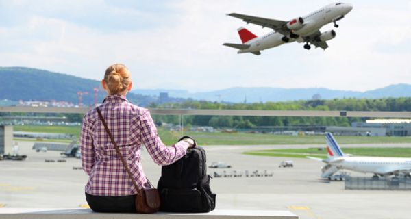 Rückansicht einer blonden Frau mit Fluggepäck in rotkarierter Bluse, die auf einer Bank sitzend den Abflug eines Flugzeuges beobachtet
