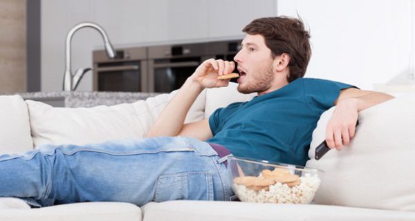 Junger Mann sitzt auf einer Couch, isst Kekse und sieht fern
