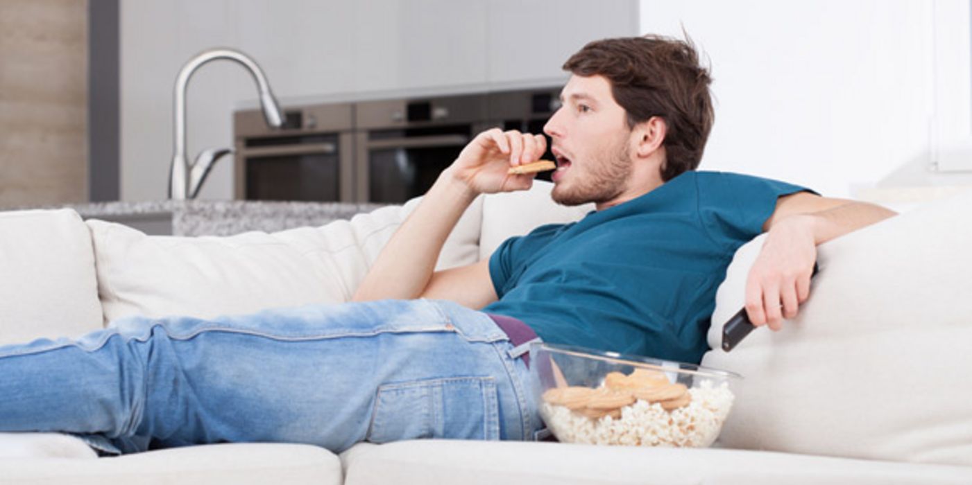 Junger Mann sitzt auf einer Couch, isst Kekse und sieht fern