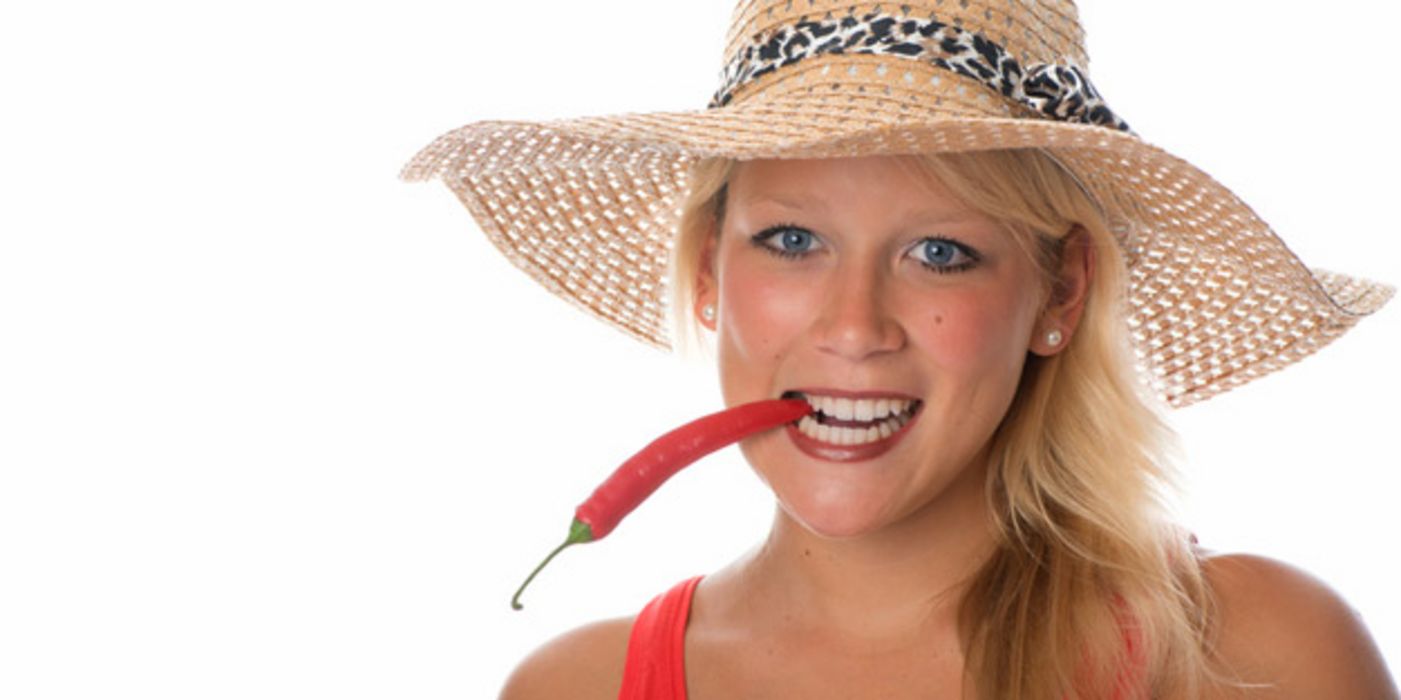 Frontalfoto sommerlich: junge Frau, rotes Trägershirt oder Kleid, Strohhut, hält rote Chilischote wie Zigarette im Mund mit den Zähnen fest
