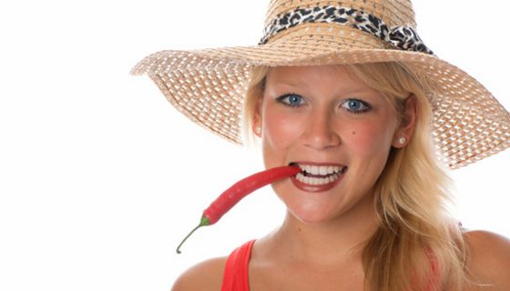 Frontalfoto sommerlich: junge Frau, rotes Trägershirt oder Kleid, Strohhut, hält rote Chilischote wie Zigarette im Mund mit den Zähnen fest