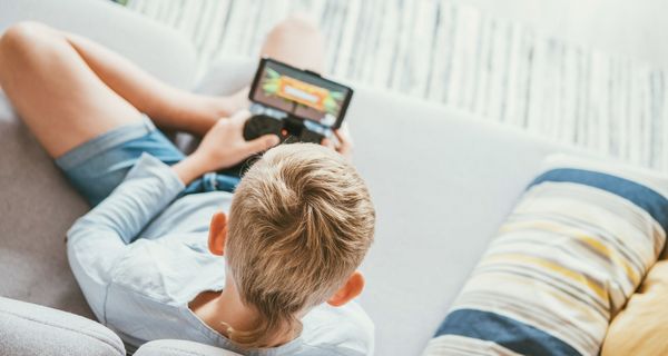 Blonder Junge, sitzt auf einer Couch und spielt mit einer Spielekonsole. 